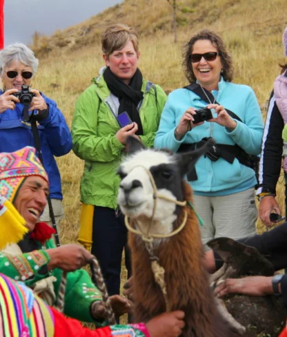 Group of people in colorful clothing gather around a llama in a field; some are taking photos.