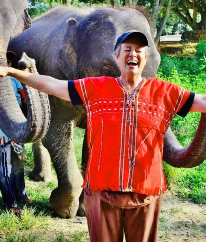 A person in a red shirt stands smiling with arms outstretched between two elephants in a grassy area.