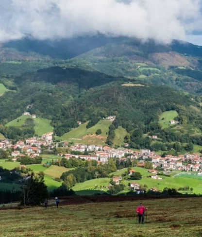 A verdant valley with a small town nestled among green hills. Three people are standing in the foreground, and mountains with clouds loom in the background.