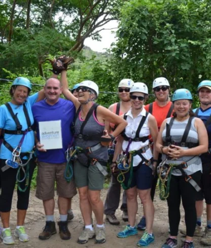 A group of people wearing helmets and harnesses stand together outdoors, preparing for a zipline adventure. They are smiling and surrounded by trees.