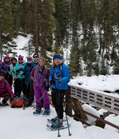 A group of people in winter gear and snowshoes stands on a snow-covered bridge surrounded by trees.