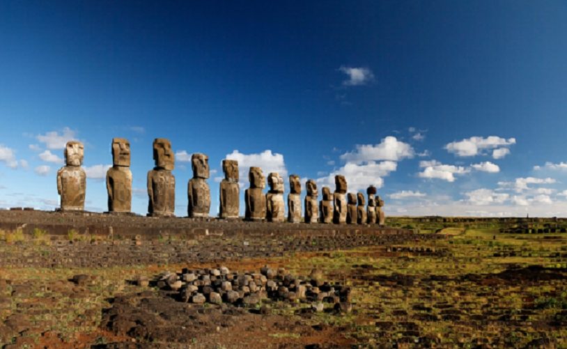 A row of large stone Moai statues stands against a blue sky on Easter Island, surrounded by a grassy landscape.