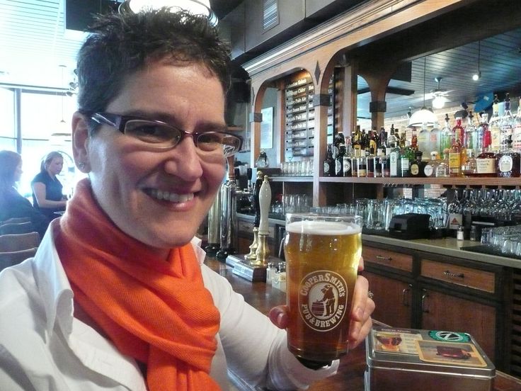 Person holding a glass of beer with a brewery logo, sitting in a bar with various bottles and taps in the background.