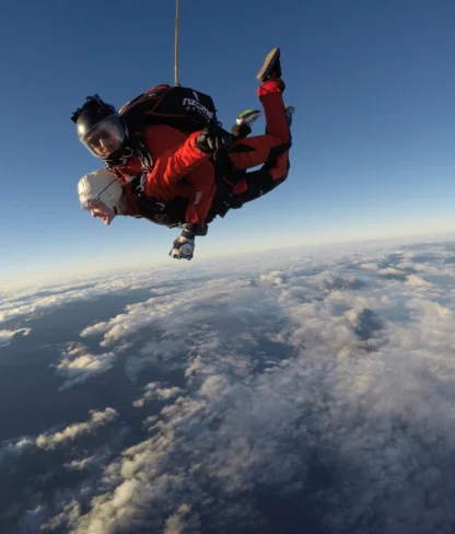 Two people tandem skydiving above the clouds under a clear blue sky.