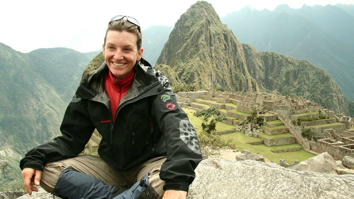 A person sits on a rock at Machu Picchu, smiling, with mountains and ancient ruins in the background.