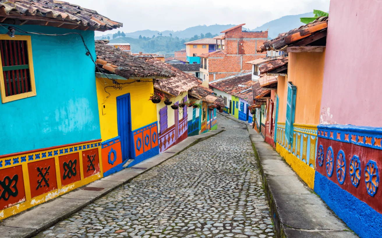 A colorful cobblestone street is lined with vibrant buildings painted in blue, yellow, orange, and green, featuring decorative patterns and wooden shutters, extending down a gentle hill.