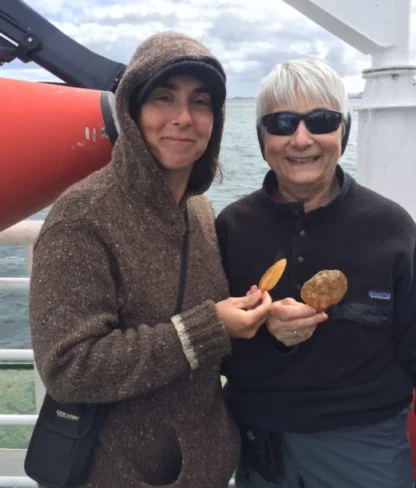 Two people in warm clothing stand on a boat deck, each holding up a potato. One wears a hood, the other sunglasses. An orange boat and ocean are in the background.