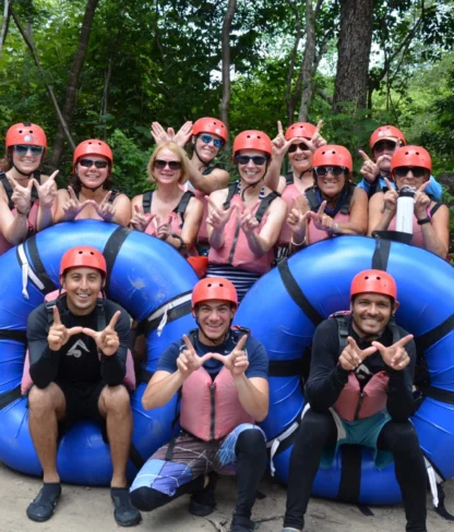 Group of people wearing helmets and life jackets posing with blue rafts in a forested area.