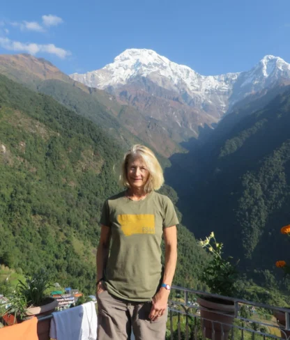 A woman stands on a balcony with mountains in the background, under a clear blue sky. Flowers are in the foreground.