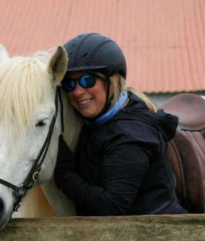 Person in a black helmet and jacket hugging a white horse over a wooden fence, with a red corrugated roof in the background.