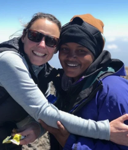 Two people in outdoor gear smile and embrace on a mountain with a clear sky in the background.