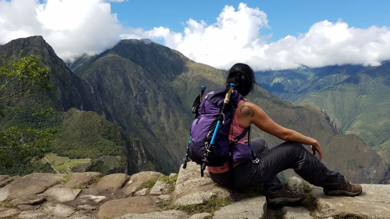 A person with a backpack sits on a rocky ledge overlooking mountainous scenery and cloudy skies.