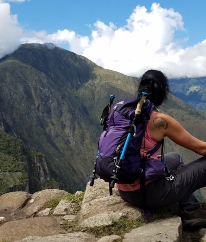 A person with a backpack sits on a rocky ledge overlooking mountainous scenery and cloudy skies.