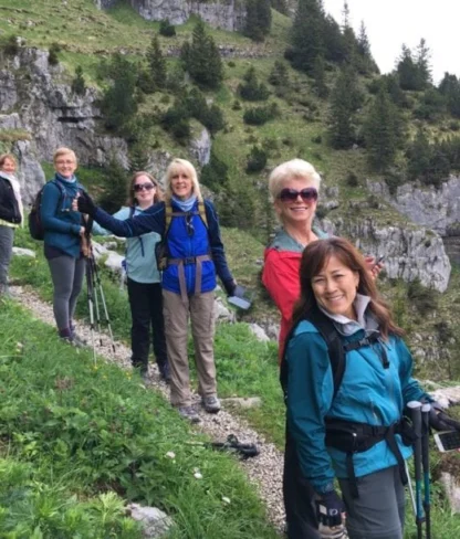 A group of six people with hiking gear on a narrow trail in a mountainous area with grass and rocks.