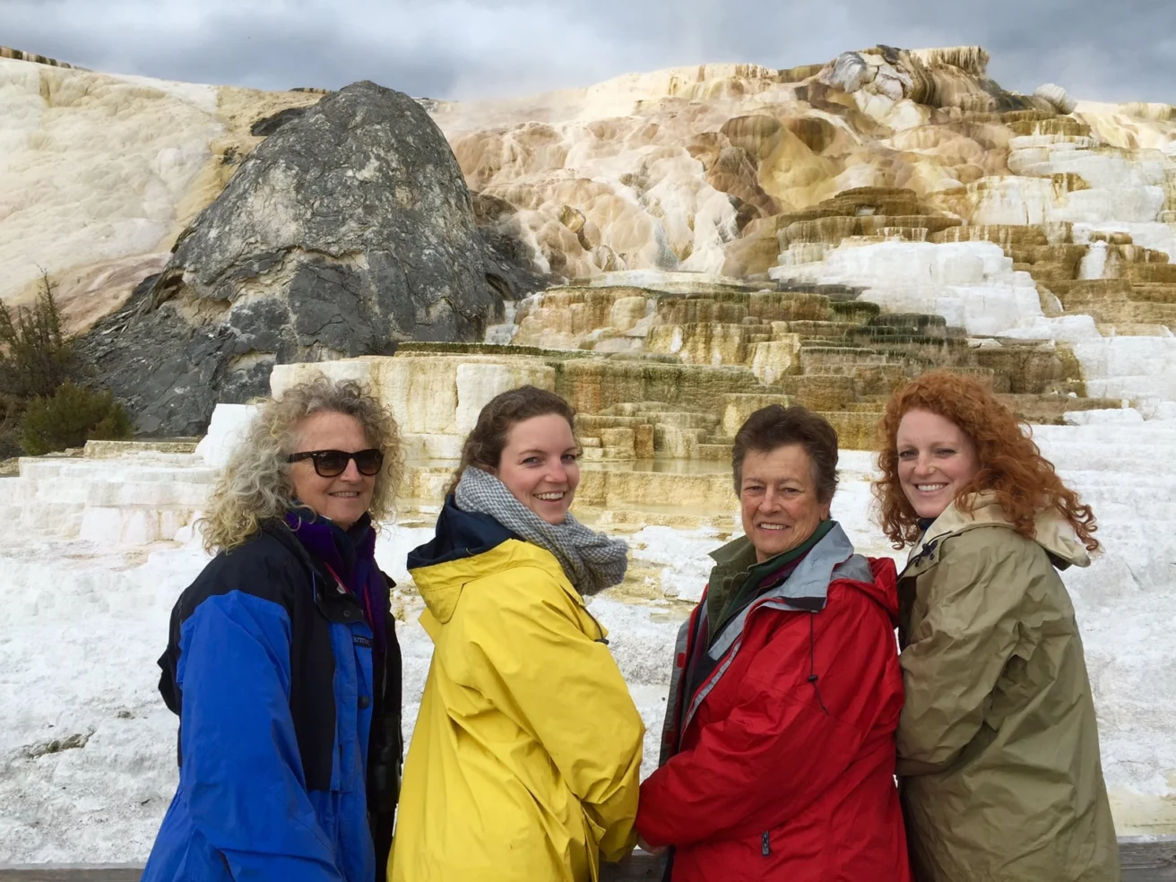 Four people in colorful jackets stand in front of layered geothermal terraces with rock formations in the background.