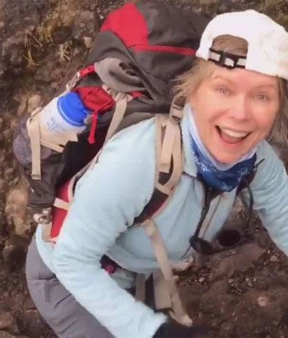 A person in outdoor gear climbs a rocky terrain, smiling at the camera. They wear a backpack, cap, and a blue shirt.