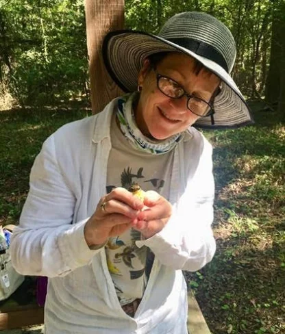 Person wearing a sunhat and glasses holds a small bird outdoors in a wooded area.