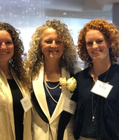 Three women with curly hair stand together indoors, wearing name badges and smiling at the camera.