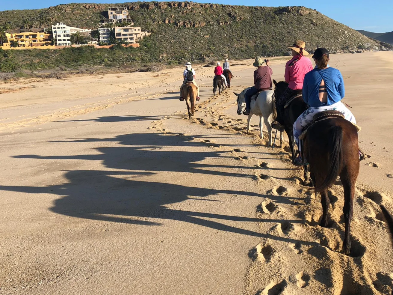 People horseback riding on a sandy beach, leaving a trail of hoofprints, with hills and buildings in the background.