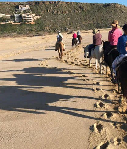 People horseback riding on a sandy beach, leaving a trail of hoofprints, with hills and buildings in the background.