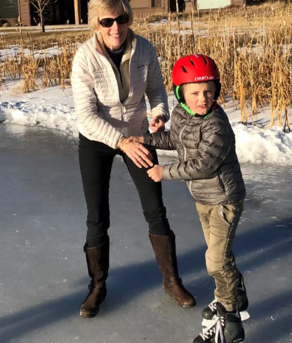 A woman and a child in a red helmet ice skating together on a frozen outdoor pond in a sunny winter setting.