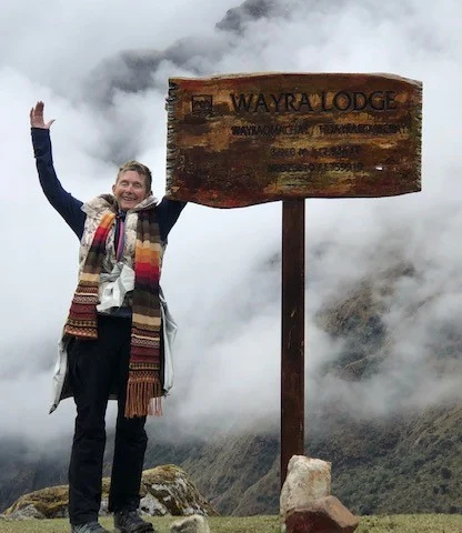 Person smiling with arms raised next to a wooden sign for Wayra Lodge, surrounded by fog and mountains.