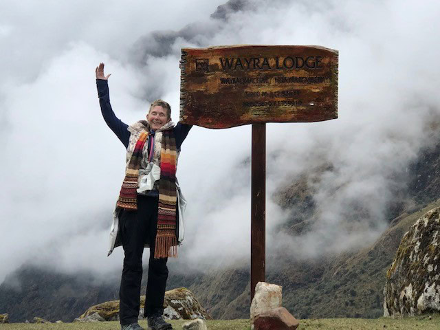 Person smiling with arms raised next to a wooden sign for Wayra Lodge, surrounded by fog and mountains.