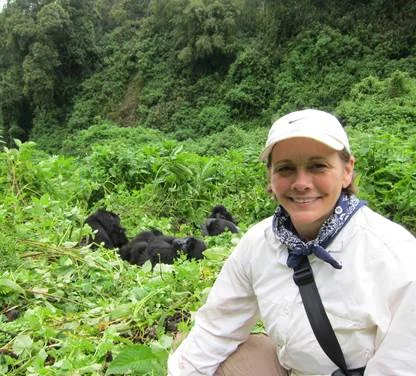 A person in outdoor attire smiles at the camera. In the background, gorillas rest among lush greenery.