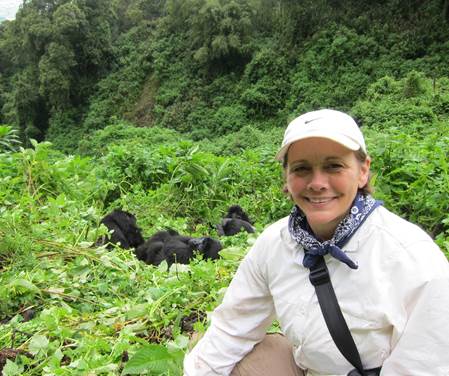 A person in outdoor attire smiles at the camera. In the background, gorillas rest among lush greenery.