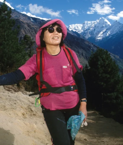 A person hiking on a mountain trail, wearing a pink hat and sunglasses, holding a walking stick. Snow-capped mountains and trees are in the background.