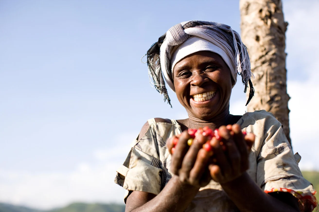 Person smiling broadly, wearing a headscarf and holding handfuls of red berries outdoors under clear sky.