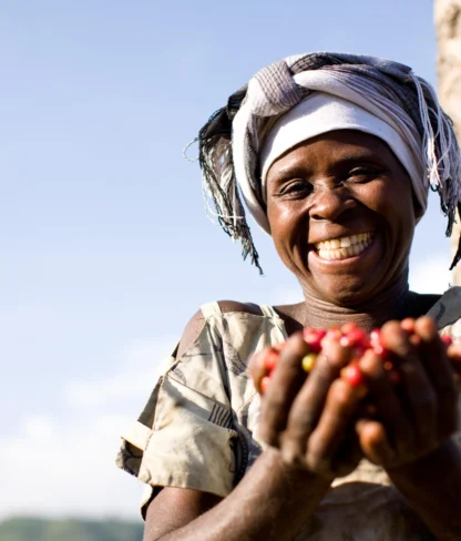 Person smiling broadly, wearing a headscarf and holding handfuls of red berries outdoors under clear sky.