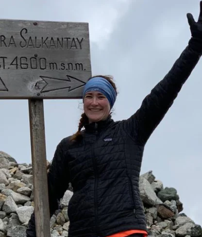 Person smiling and waving next to a sign reading "Abra Salkantay Alt. 4600 m.s.n.m." with a cloudy sky in the background.