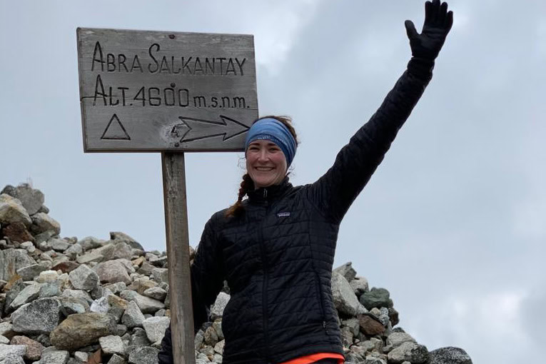 Person smiling and waving next to a sign reading "Abra Salkantay Alt. 4600 m.s.n.m." with a cloudy sky in the background.