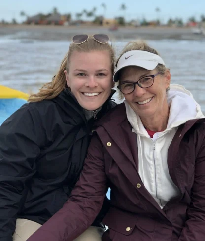 Two people smiling on a boat with a river and distant shoreline in the background.