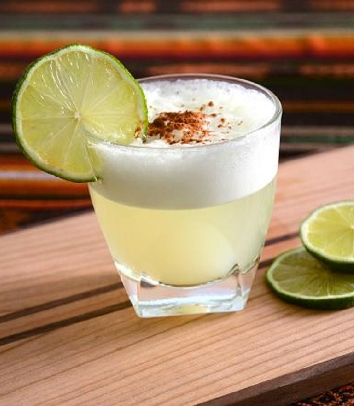 A glass of frothy yellow cocktail garnished with a lime slice and sprinkled spice, placed on a wooden board with lime slices beside it.