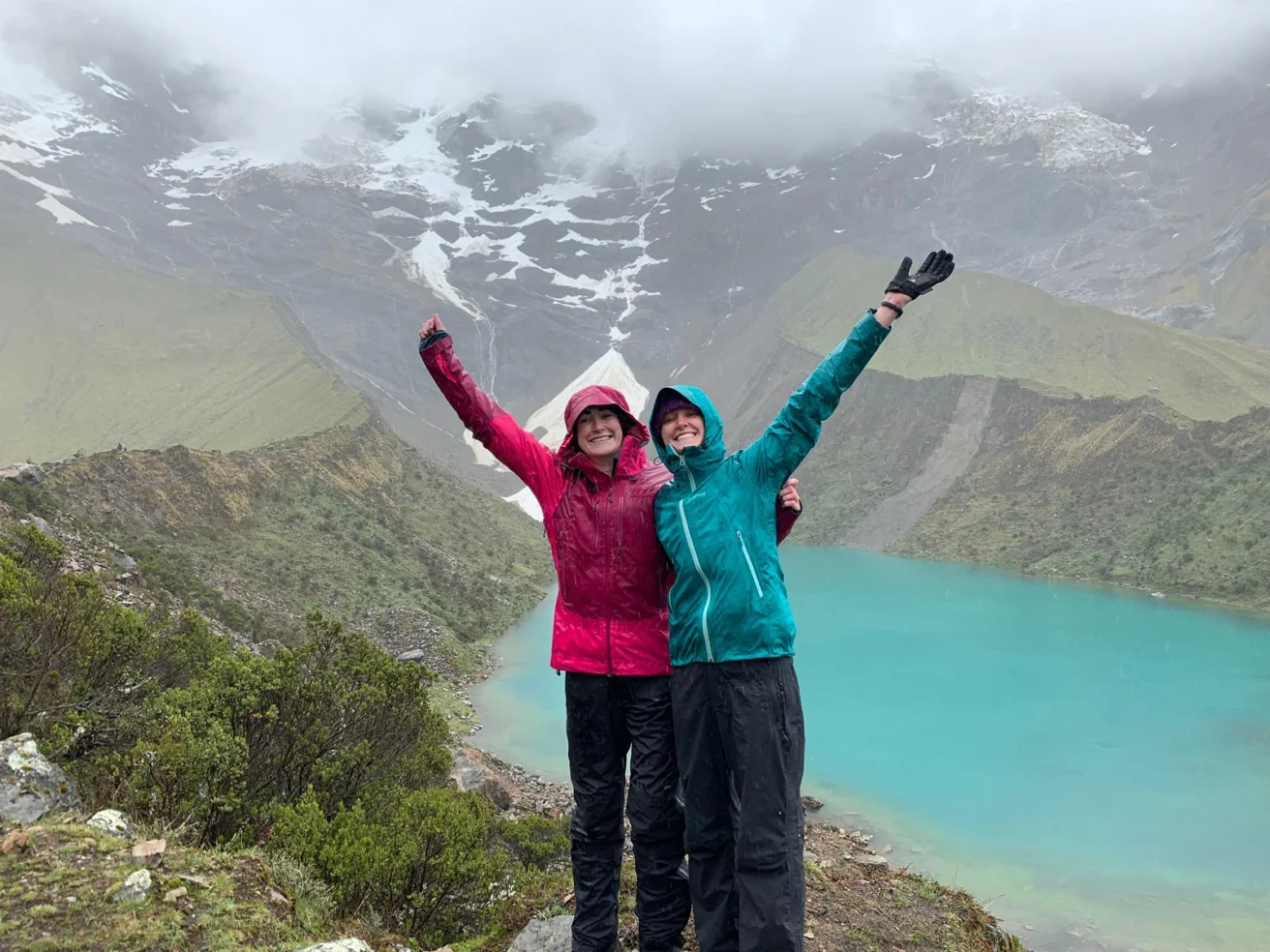 Two people in colorful jackets stand with arms raised near a turquoise mountain lake, surrounded by lush greenery and misty, snow-capped peaks.