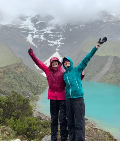 Two people in colorful jackets stand with arms raised near a turquoise mountain lake, surrounded by lush greenery and misty, snow-capped peaks.