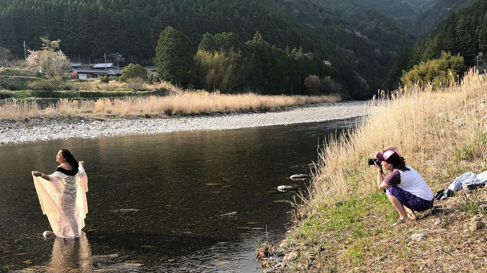 A woman stands in a shallow river holding sheer fabric while another person kneels at the riverbank, taking photos. Trees and a house are visible in the background.