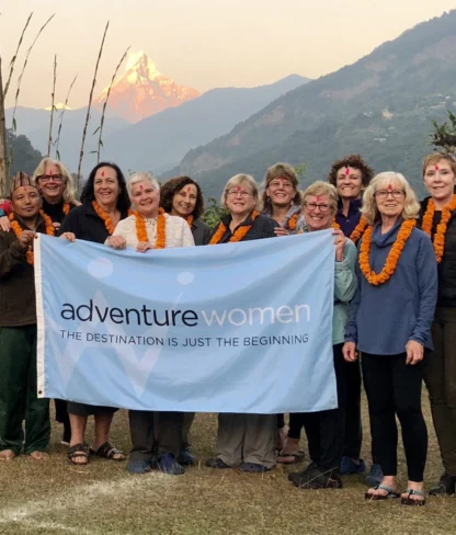 A group of women wearing orange marigold garlands hold an "Adventure Women" banner outdoors with a mountain in the background.