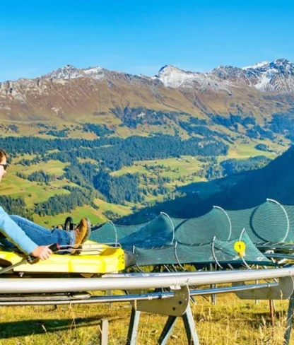 A person rides a yellow alpine coaster through mountainous terrain under a clear blue sky.