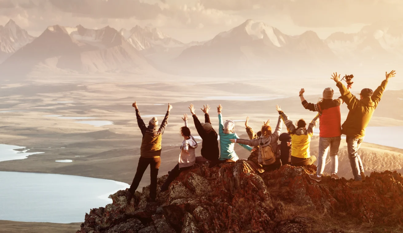 A group of people sit on a rocky cliff overlooking a vast landscape of lakes and mountains, raising their arms in celebration.