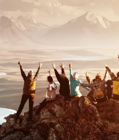 A group of people sit on a rocky cliff overlooking a vast landscape of lakes and mountains, raising their arms in celebration.