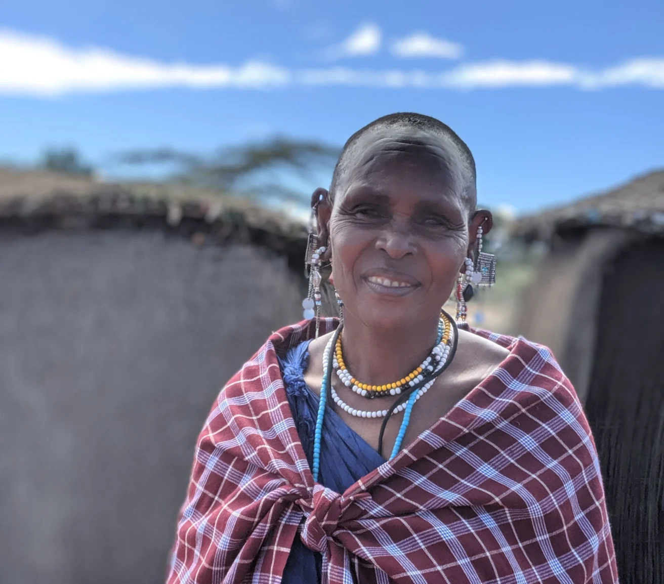 A person wearing traditional beaded jewelry and a red patterned shawl stands outdoors with huts in the background under a blue sky.