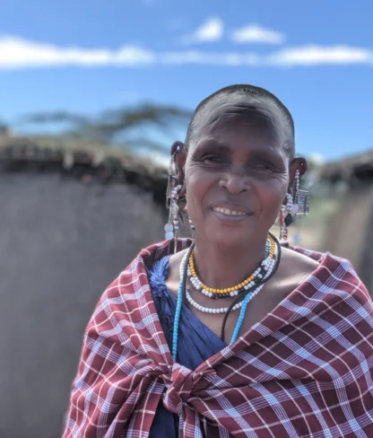 A person wearing traditional beaded jewelry and a red patterned shawl stands outdoors with huts in the background under a blue sky.