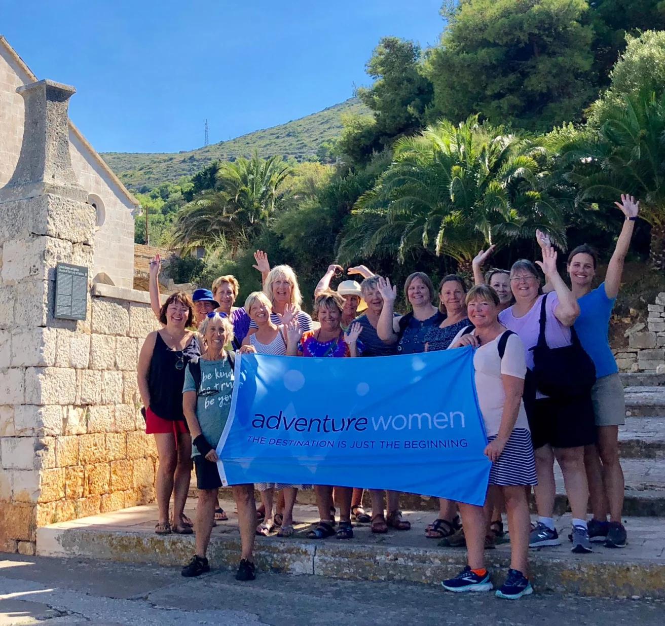 A group of women stands on stone steps outdoors, holding a blue banner that reads "adventure women: the destination is just the beginning." Trees and a hill are in the background.