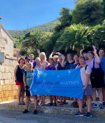 A group of women stands on stone steps outdoors, holding a blue banner that reads "adventure women: the destination is just the beginning." Trees and a hill are in the background.