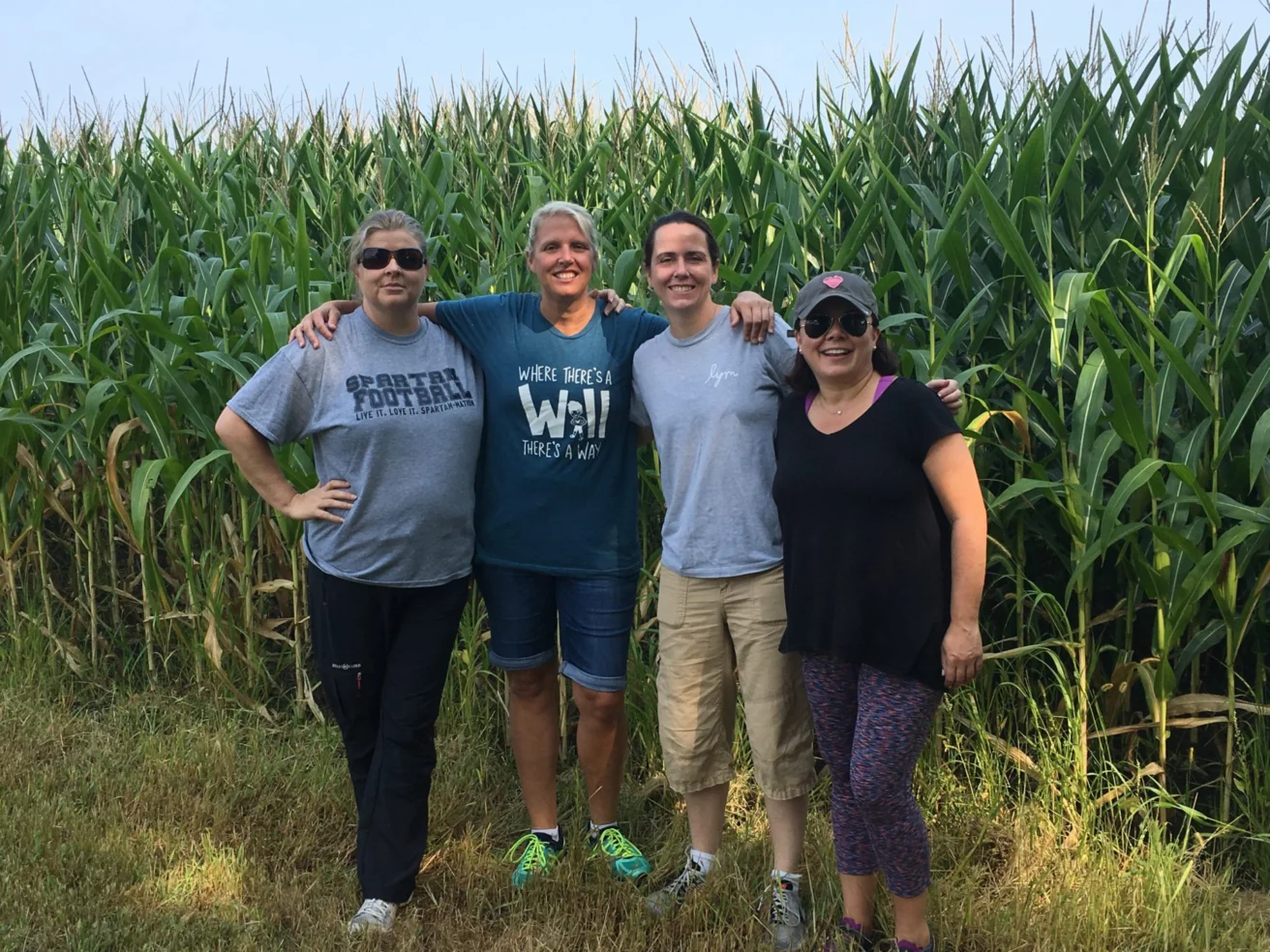 Four people stand together in front of a cornfield, smiling and posing with arms around each other. They are casually dressed and appear to be enjoying a sunny day outdoors.