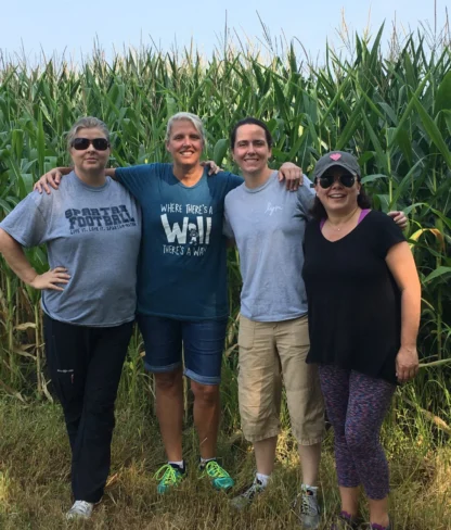 Four people stand together in front of a cornfield, smiling and posing with arms around each other. They are casually dressed and appear to be enjoying a sunny day outdoors.