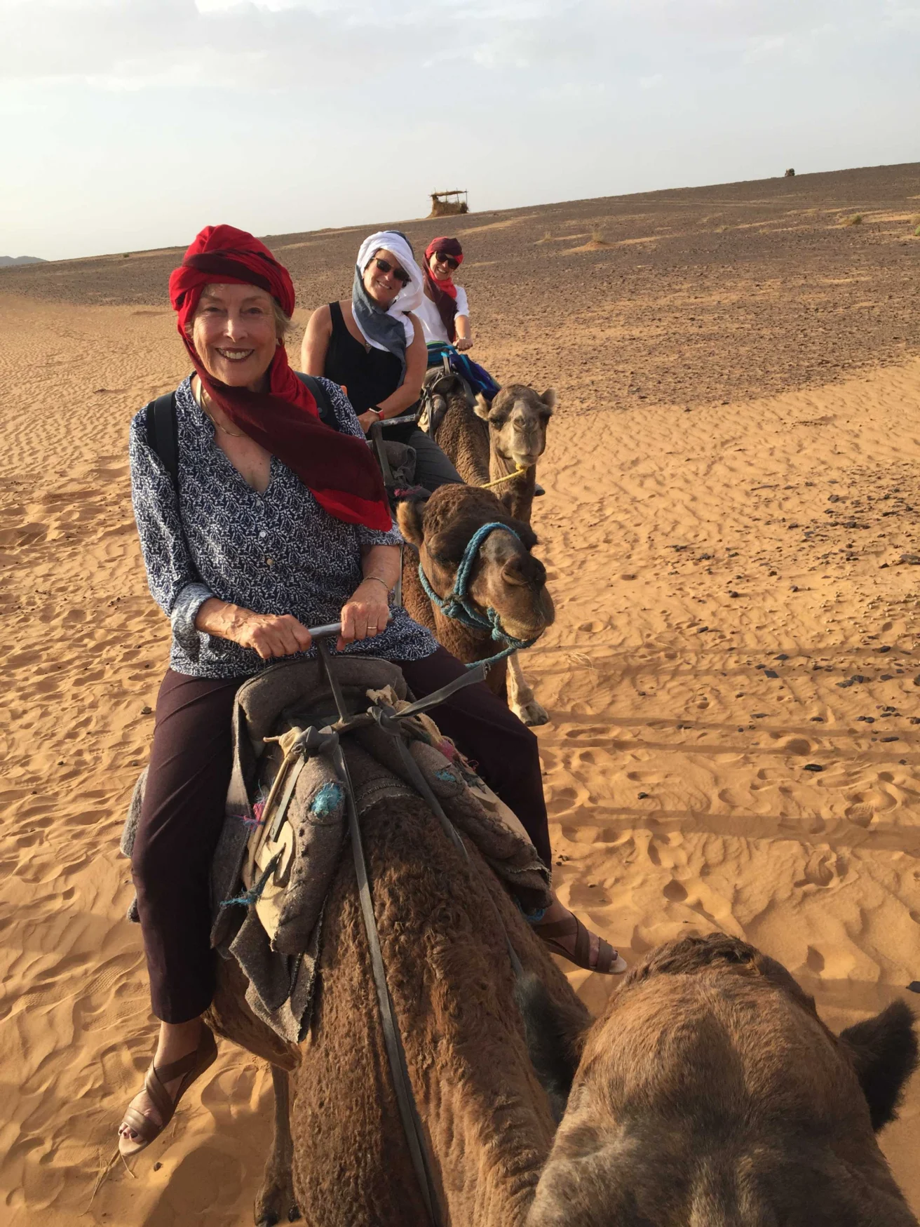 Three people riding camels in a desert landscape, wearing headscarves and casual attire under a clear sky.